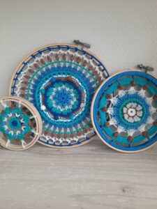 Drie gehaakte mandala's in borduurringen aan de muur, in blauw- en beigetinten