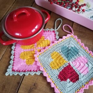Twee gehaakte pannenlappen in roze, blauw en geel naast een rode pan